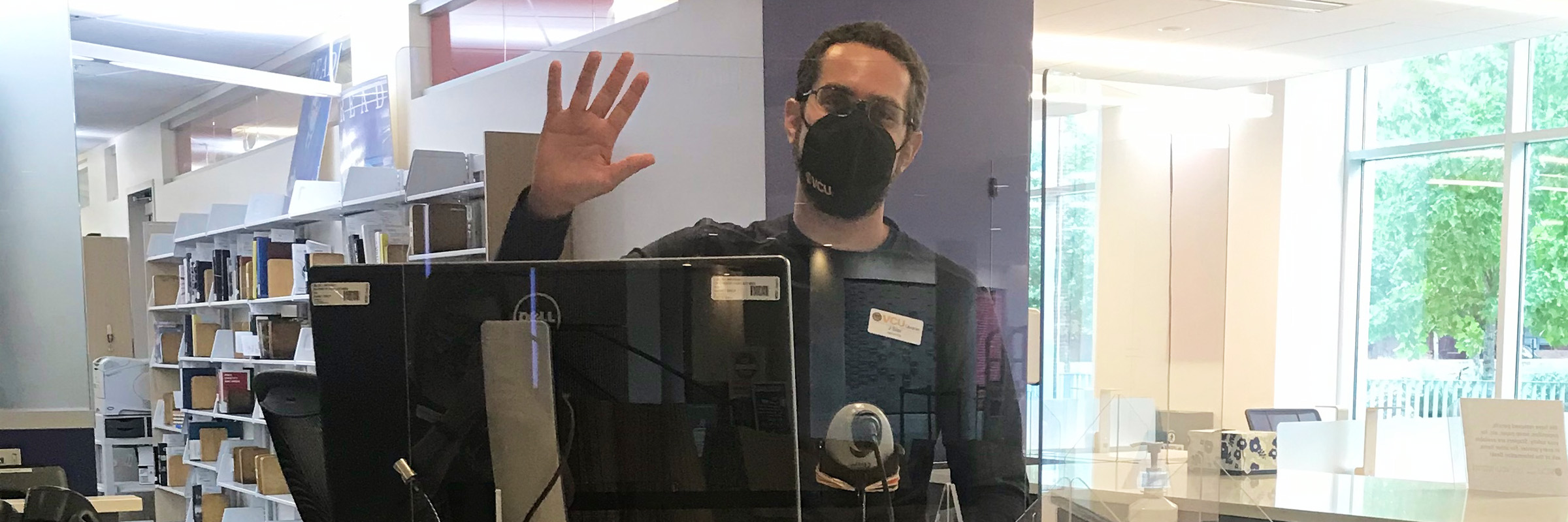 Library staff member, wearing a mask, waves behind a plexiglass shield at an information desk.